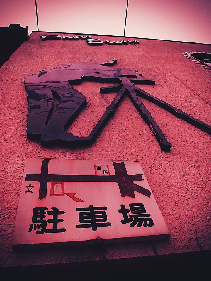 Raised dark Japanese characters cover a pink wall above a pale parking sign with a map graphic.