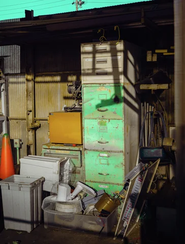 A tall green drawer cabinet stands among white containers, leaning grilles, and stored tools in a shed.