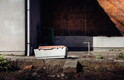 A white rectangular basin rests on a concrete ledge beside exposed taps and a boarded wall.