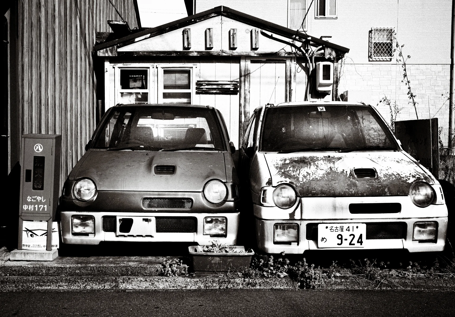 Two small weathered cars stand tightly side by side before a modest building in black and white.