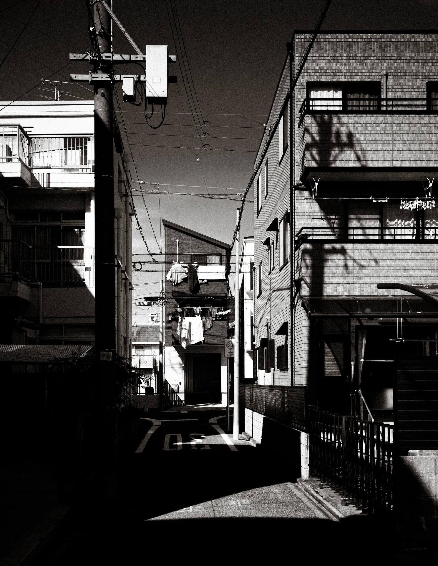 Sunlight illuminates a narrow residential alley between dark buildings and projecting balconies in black and white.