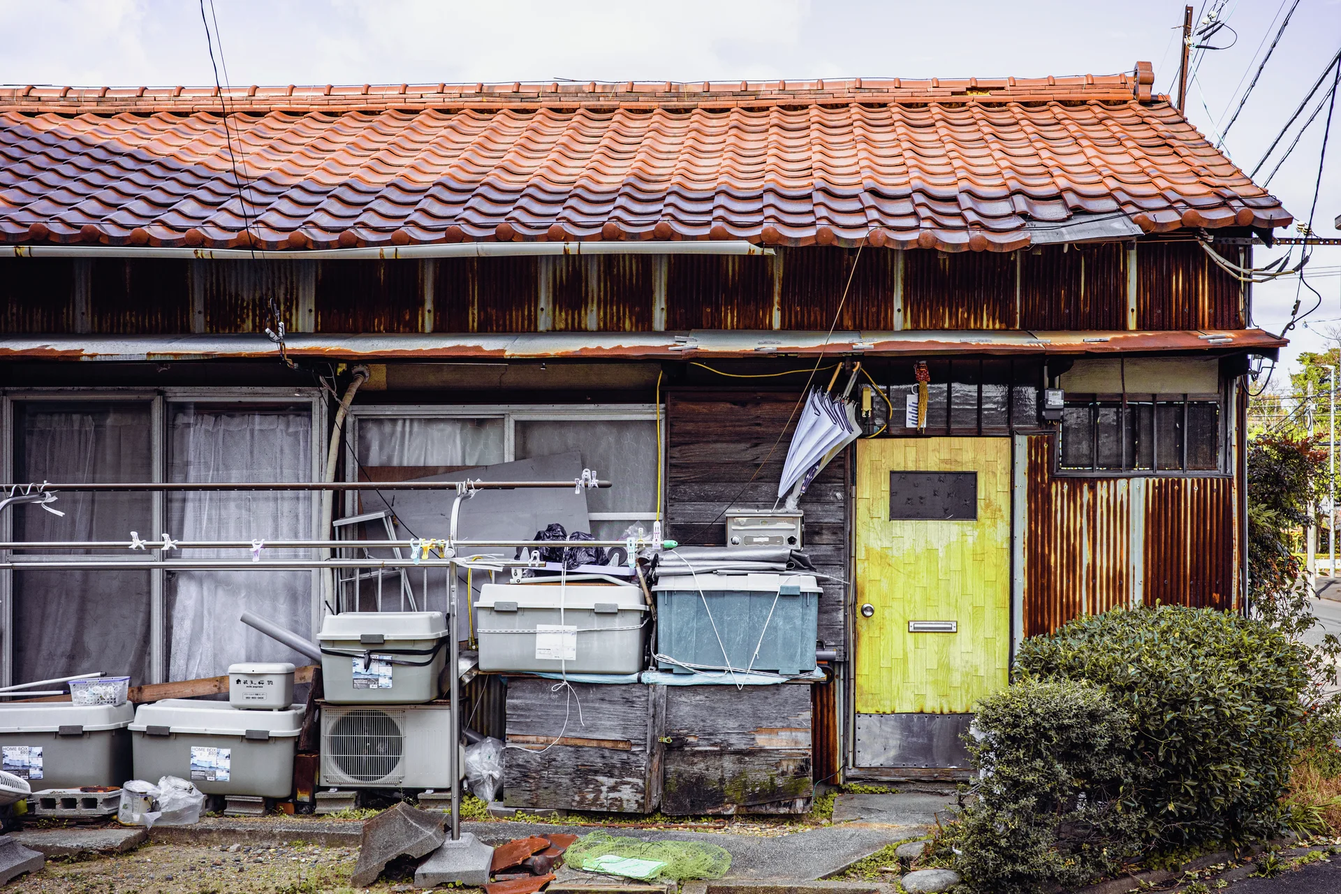 A weathered wooden house has a red tiled roof, yellow door, and assorted containers along its front.