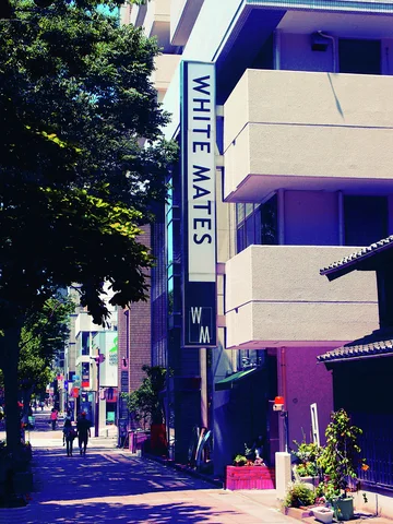 A vertical WHITE MATES sign hangs beside pale balconies above a shaded sidewalk lined with trees and pots.
