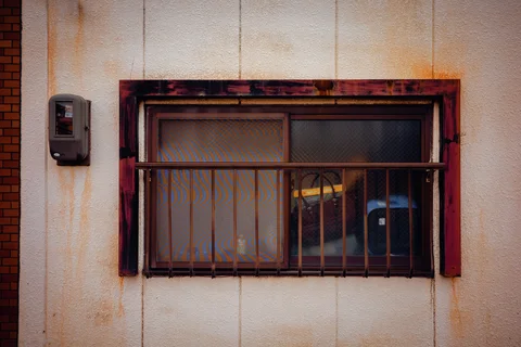 Rust stains surround a barred sliding window set in a pale wall beside an electrical box.