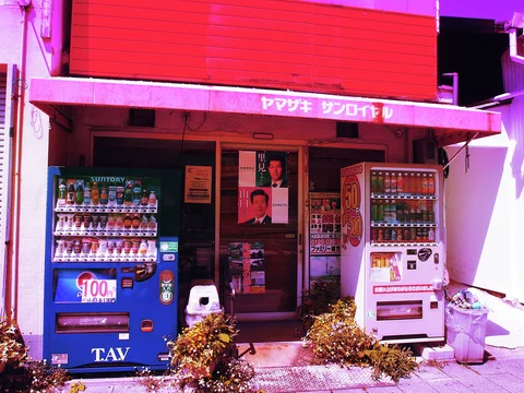 Blue and white vending machines flank a small shop entrance beneath a vivid pink awning.