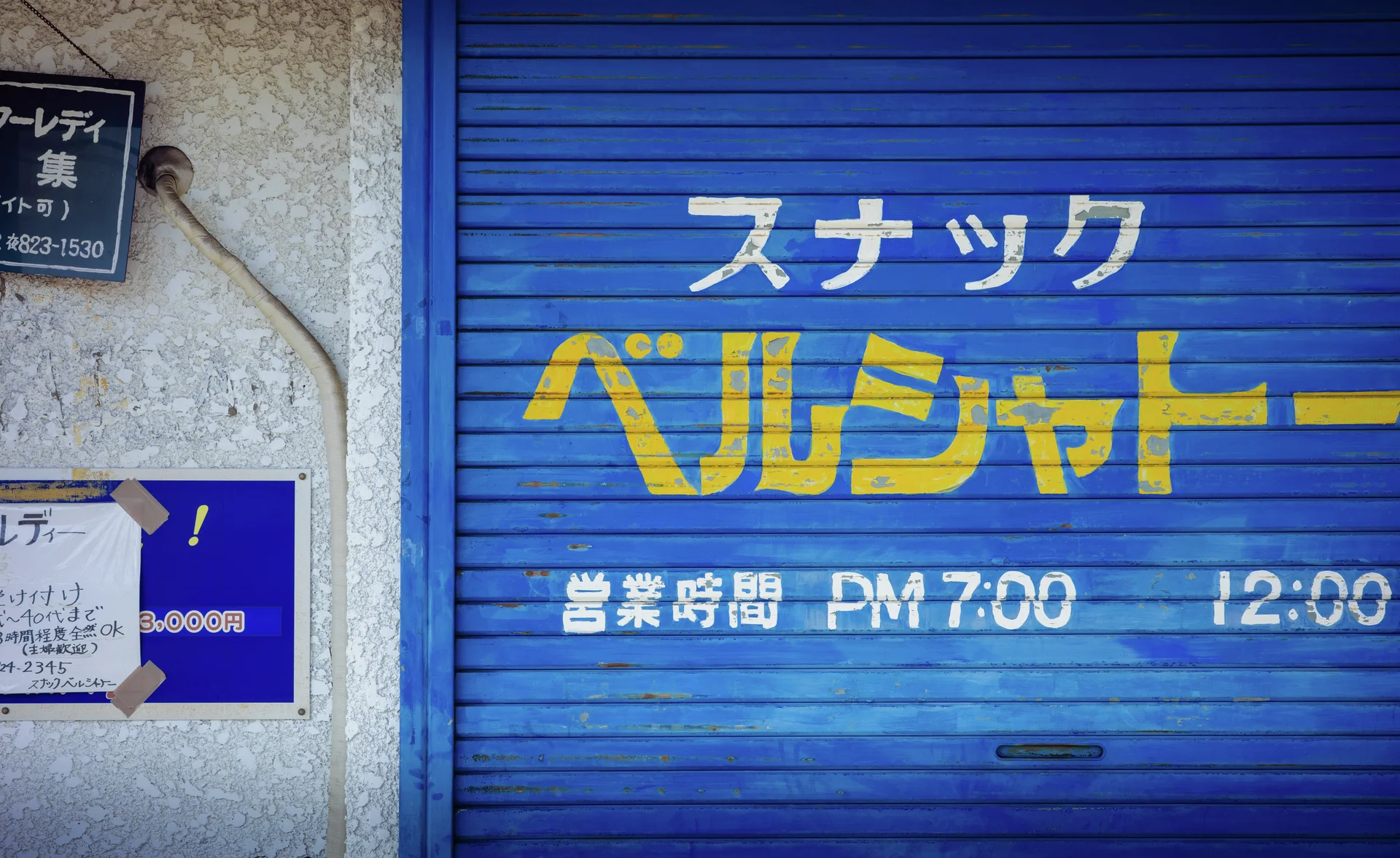 White and yellow Japanese lettering spans a closed blue metal shutter beside small wall signs.