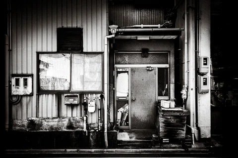 A worn narrow door sits among pipes, utility meters, and pale windows on a corrugated facade.