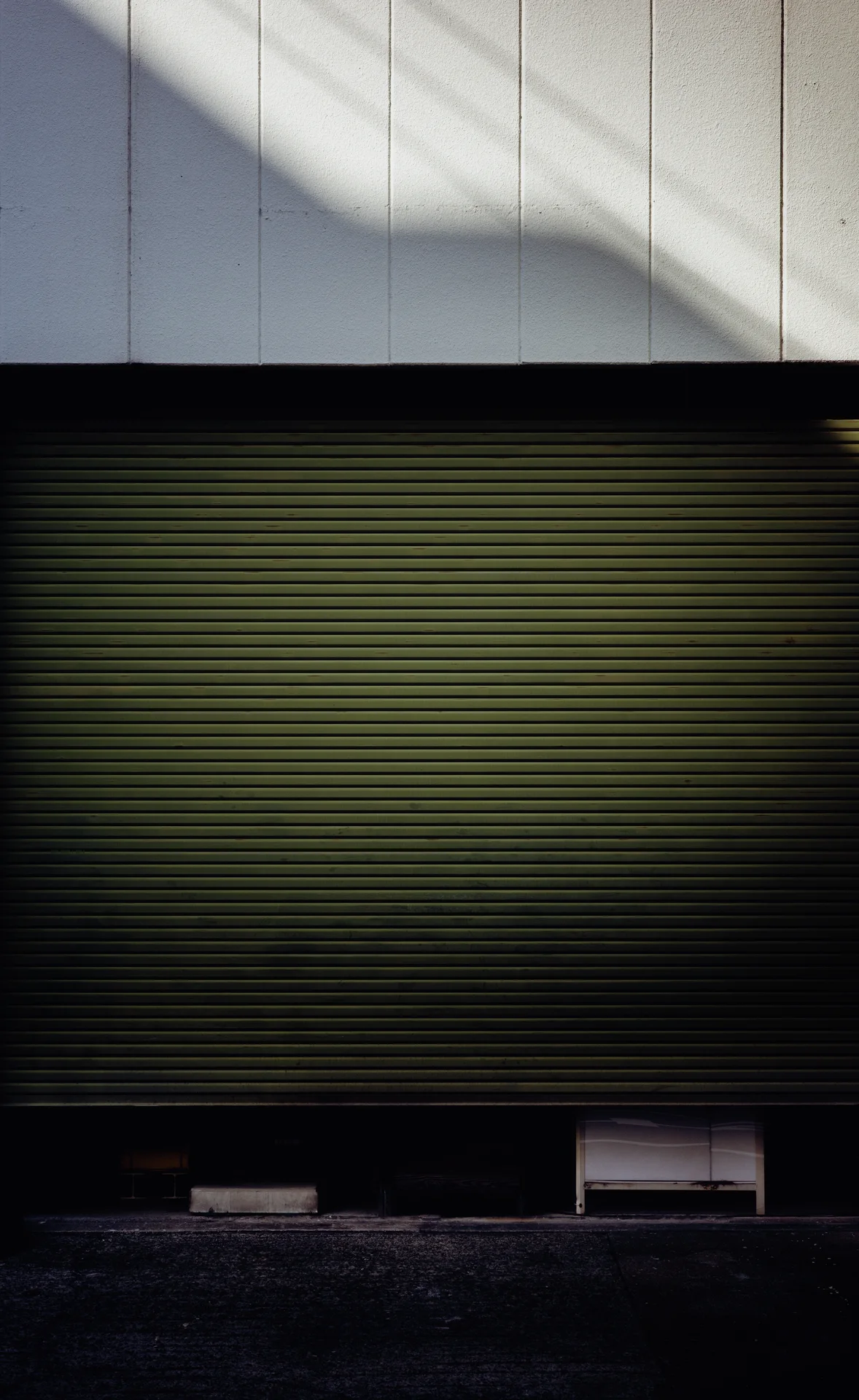 A diagonal shadow crosses pale upper panels above a closed green shutter and a dark pavement edge.