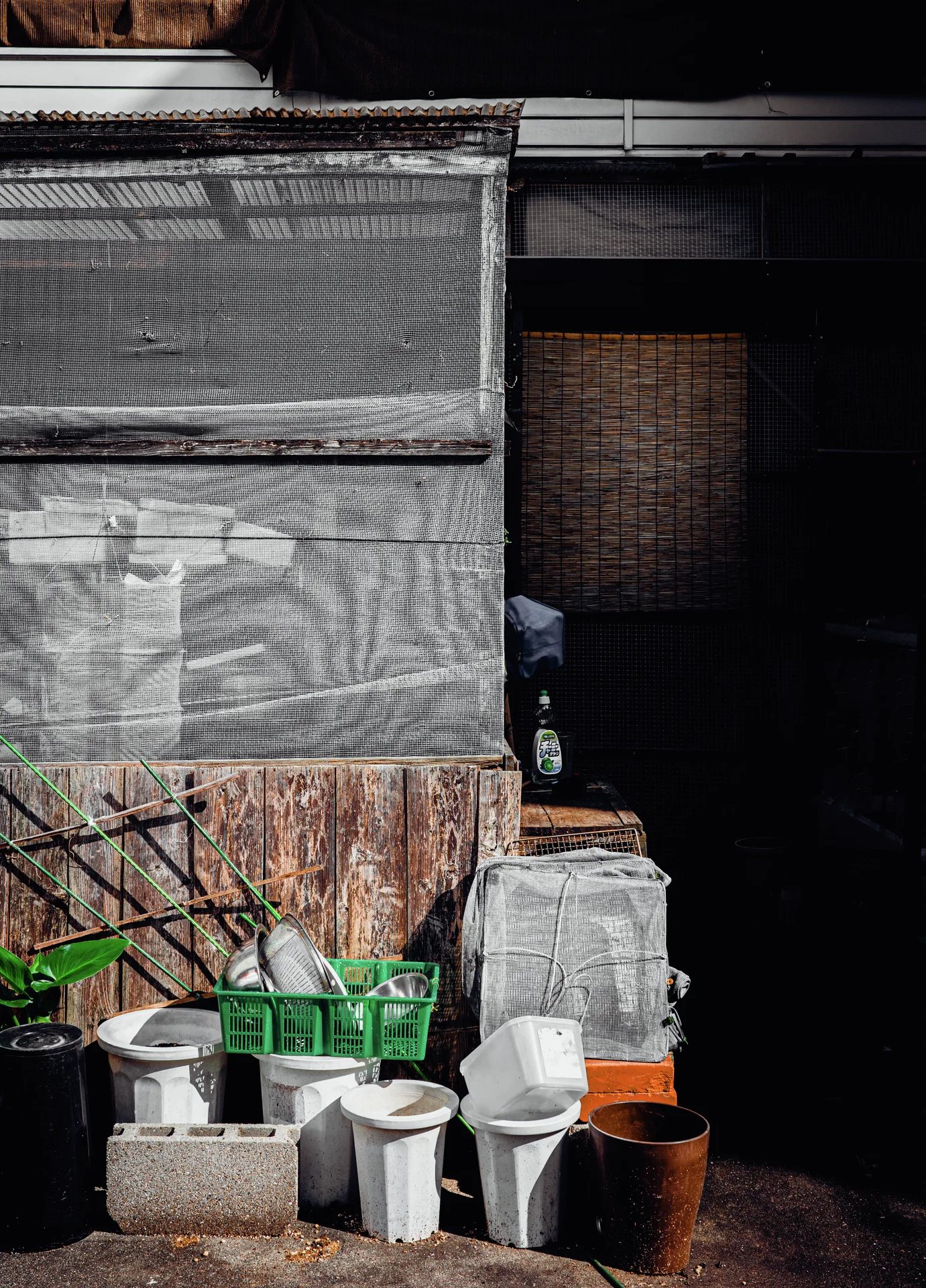 Buckets, a green crate, and concrete blocks cluster against weathered wooden boards beside a dark doorway.