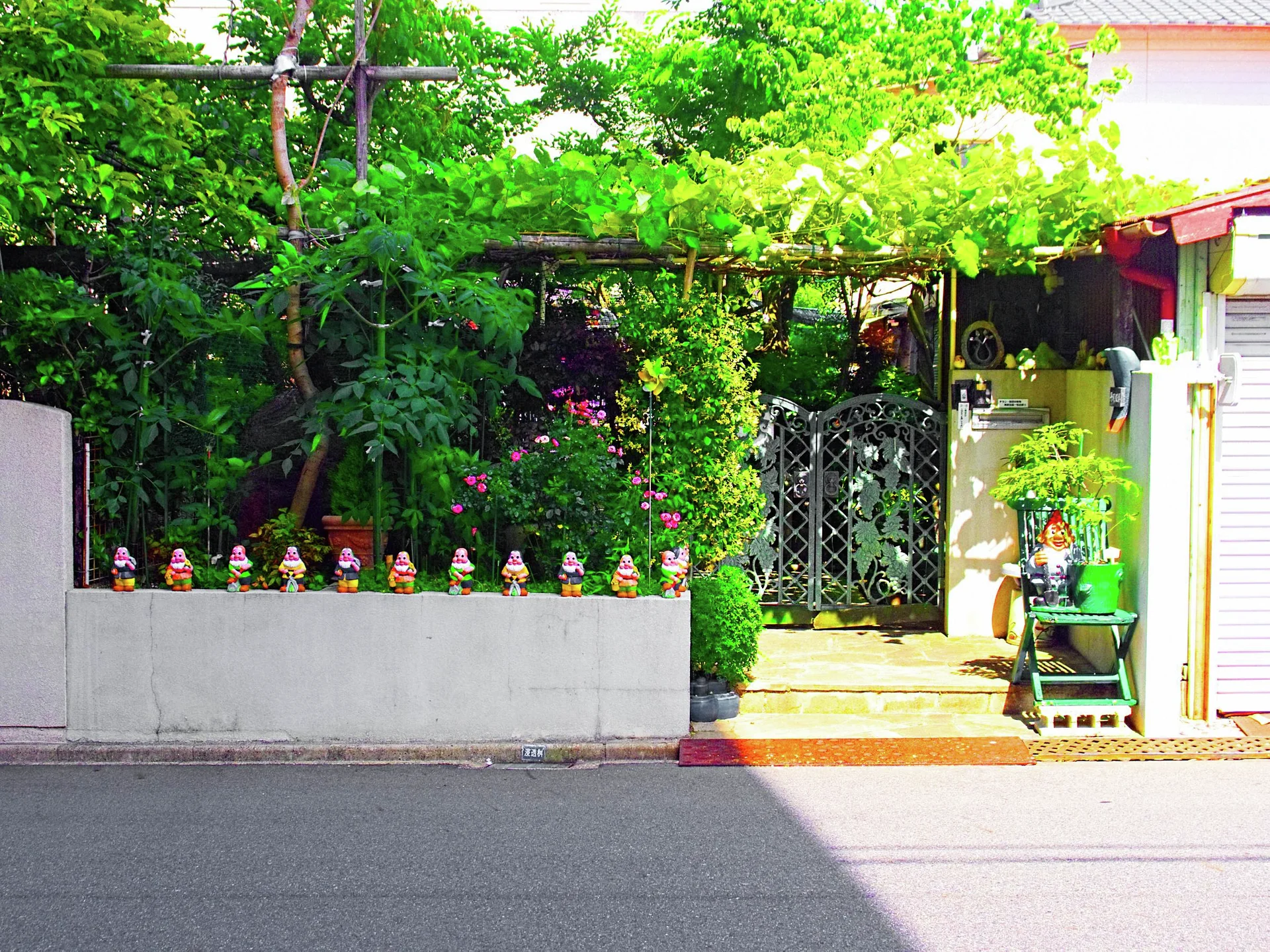 Small colorful figurines line a low wall before a leafy garden entrance with a patterned metal gate.