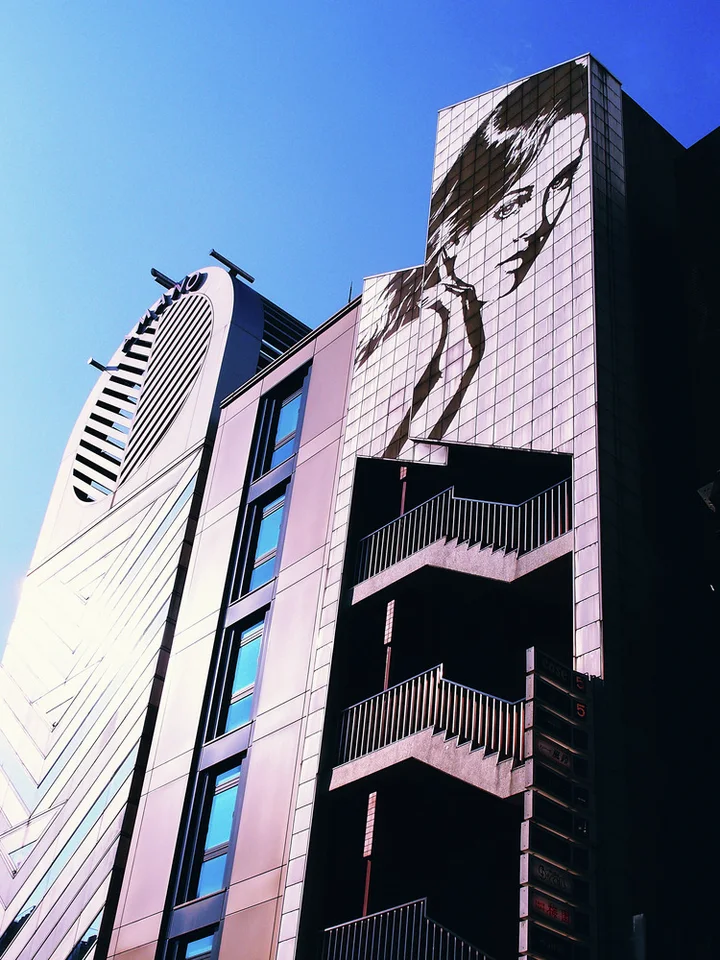 A large monochrome woman's face spans a tiled building facade beside dark exterior balconies under blue sky.