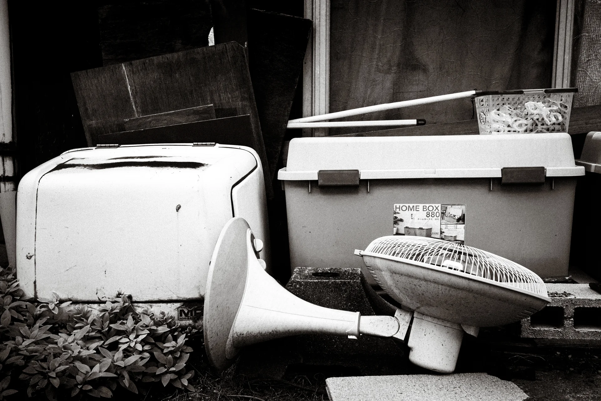 Discarded appliances and a plastic storage box crowd a shaded corner in black and white.
