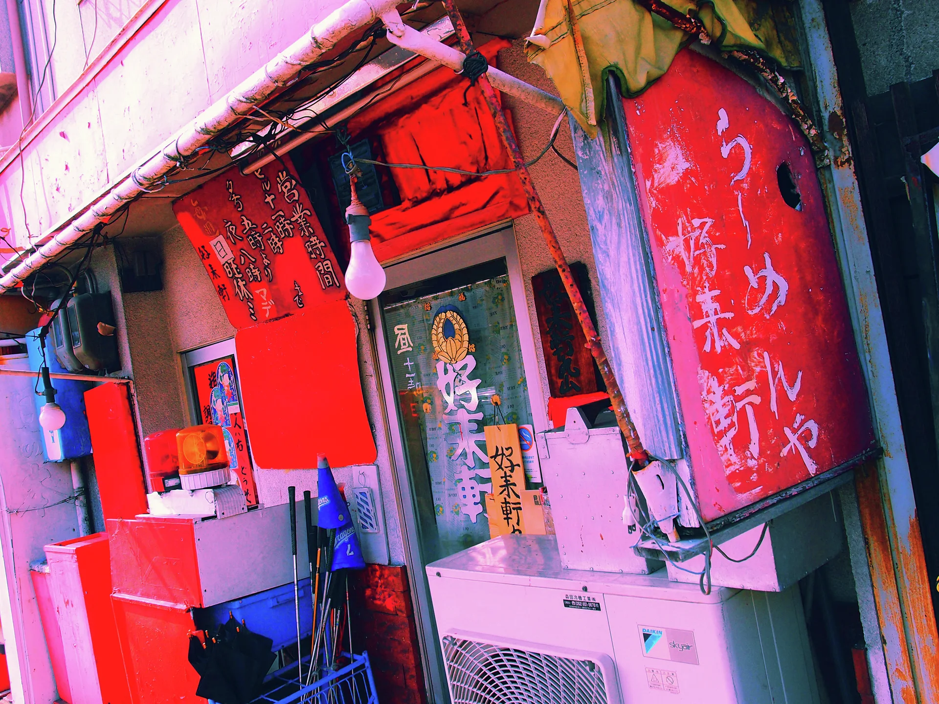 Red Japanese lanterns hang beside a narrow shop doorway above an outdoor air conditioner.