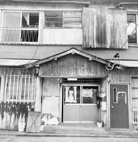 A weathered two-story facade has broken upper windows and a small gabled entrance in black and white.