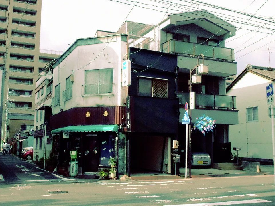 A weathered corner shop with a green awning stands beside a dark building with stacked balconies.