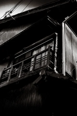 An old wooden balcony with metal railings projects from a shadowed house in monochrome.