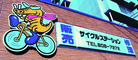 A colorful cartoon dragon rides a turquoise bicycle beside a rectangular shop sign on a brick wall.