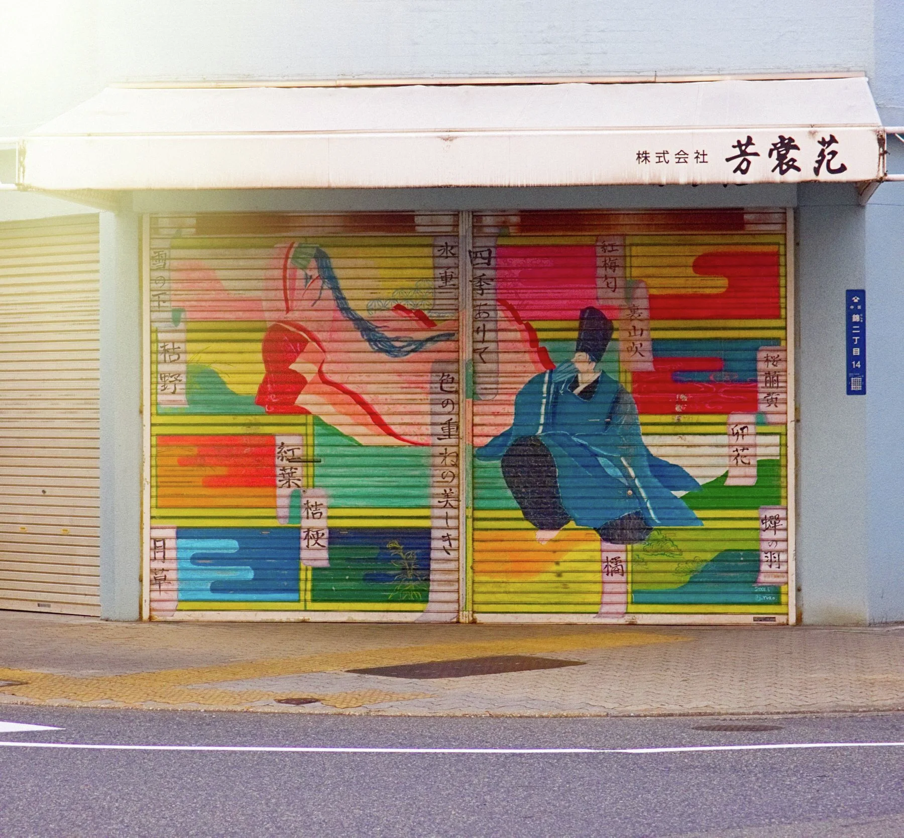 Colorful illustrated figures cover closed shop shutters beneath a pale awning and Japanese lettering.