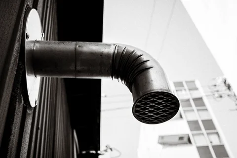 A curved metal vent pipe projects from a wall, ending in a diamond-patterned grille.
