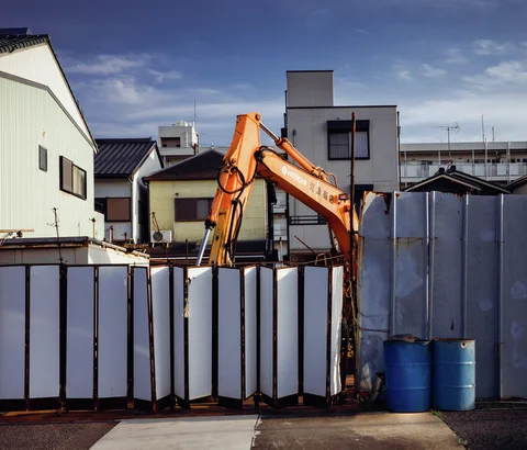 An orange excavator rises behind pale construction panels beside two blue barrels and small buildings.