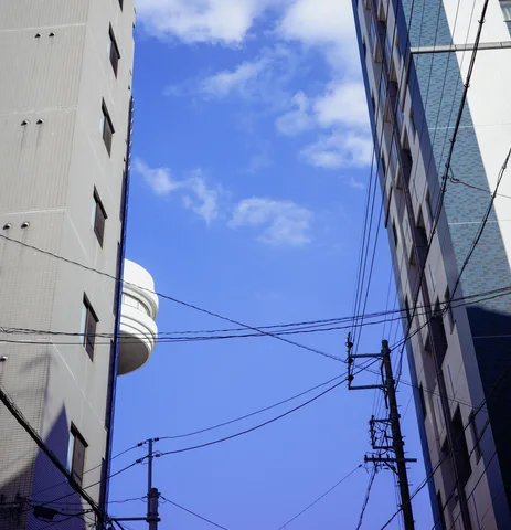 Overhead cables crisscross a blue strip of sky framed by tall pale building walls.