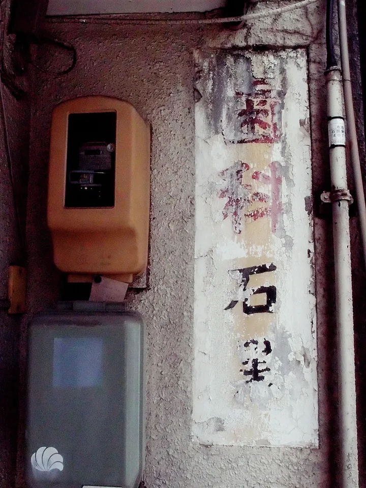 Faded vertical Japanese lettering remains on a stained wall beside two old electrical meter boxes.