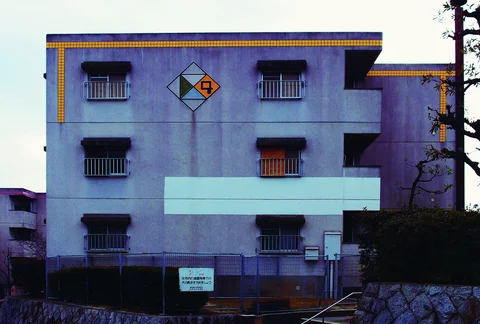 A pale apartment facade has rows of barred windows, yellow roof trim, and a colored diamond emblem.