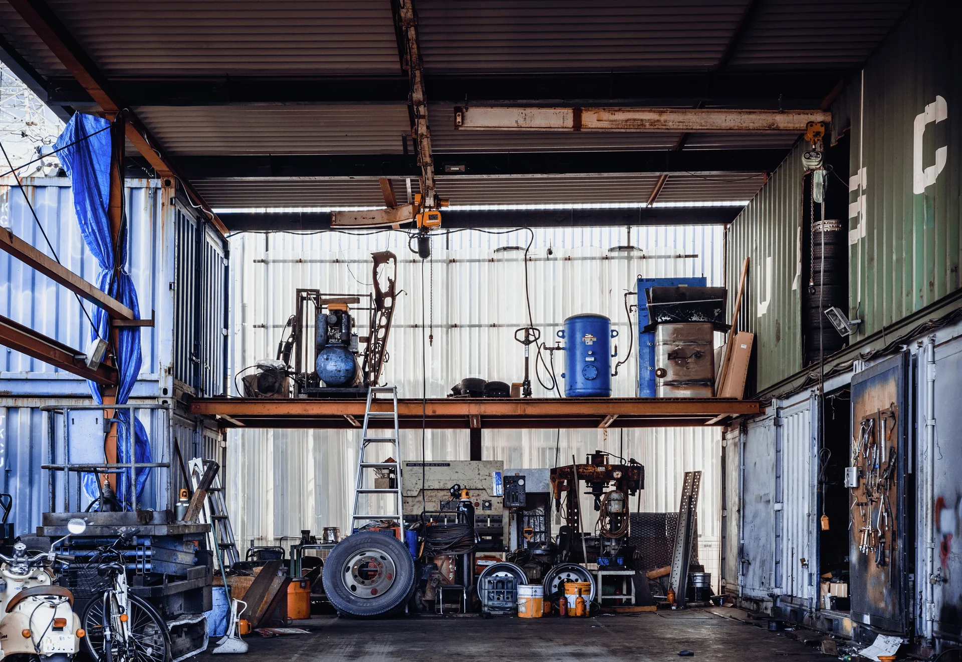 Tools, containers and vehicle parts occupy two levels of an open corrugated metal workshop.