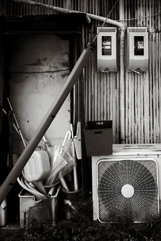 Meters, bundled pipes, and an air conditioner cluster beside corrugated siding and a diagonal brace in black and white.