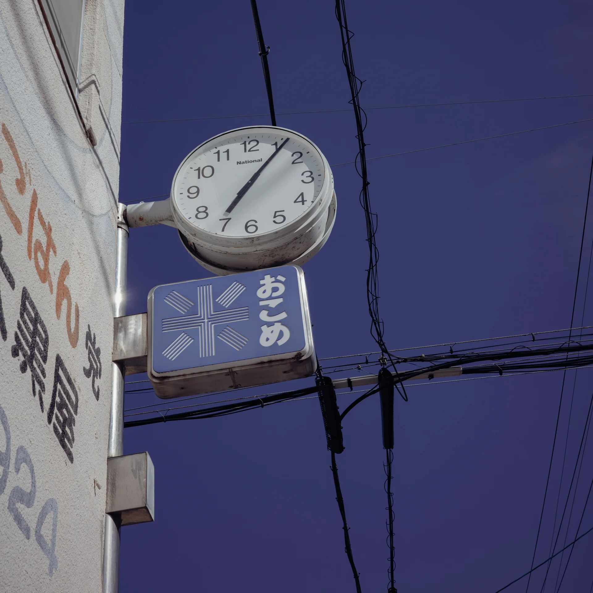 A round clock projects above a blue shop sign against a sky crossed by wires.