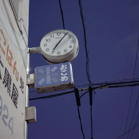 A round clock projects above a blue shop sign against a sky crossed by wires.