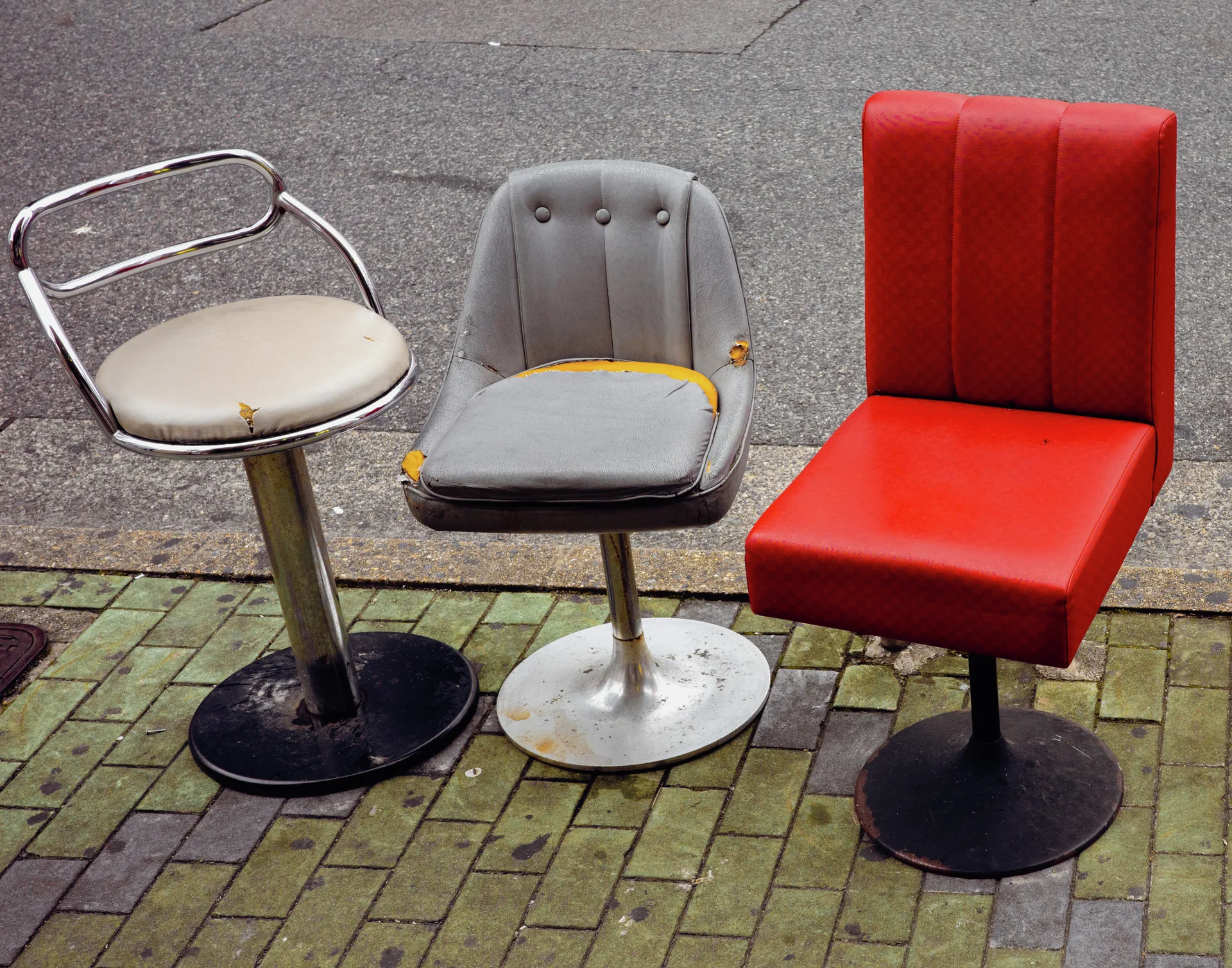 Three mismatched pedestal chairs, including a bright red seat, stand on a brick sidewalk.