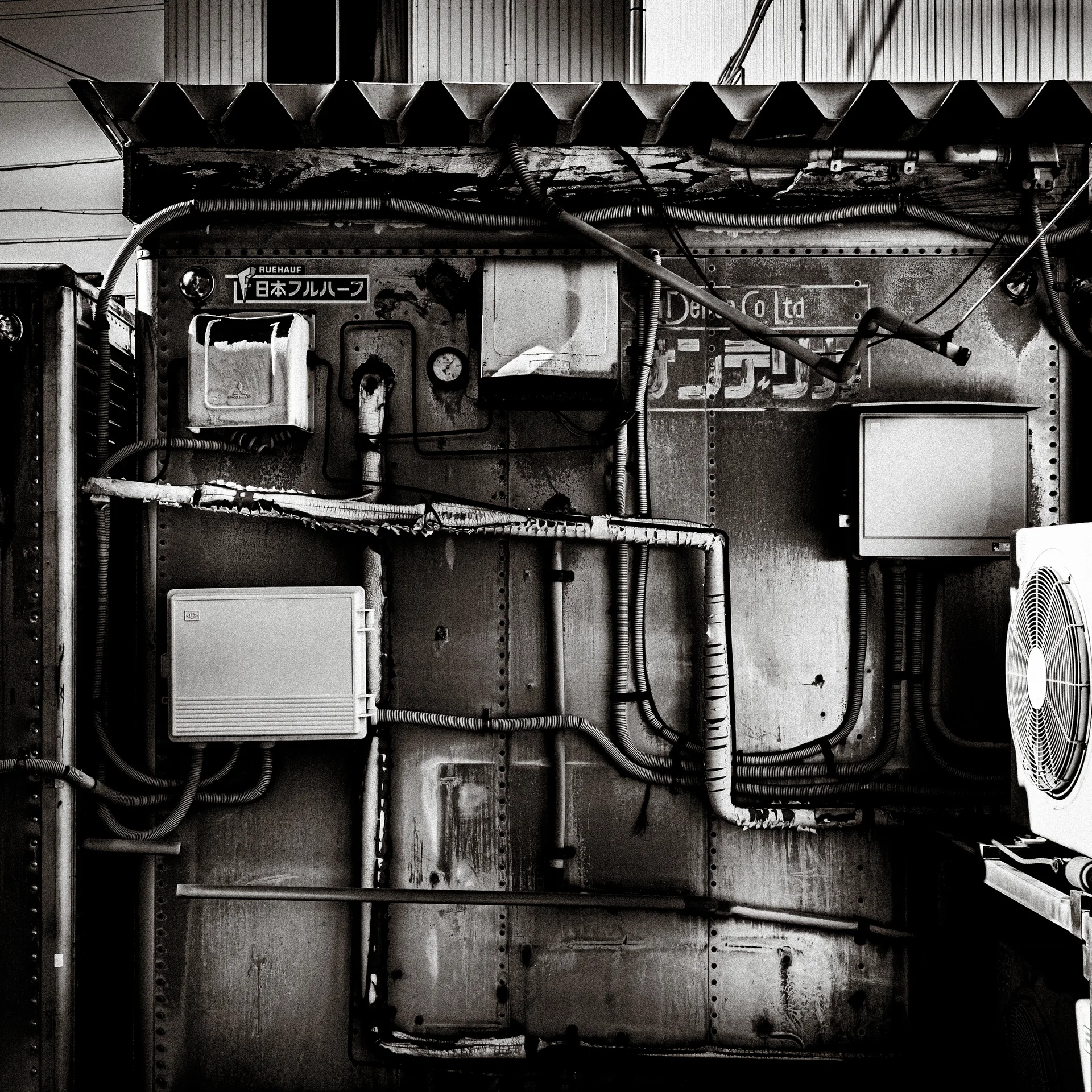 Meters, boxes, and tangled cables cover patched metal siding beneath a corrugated awning in black and white.