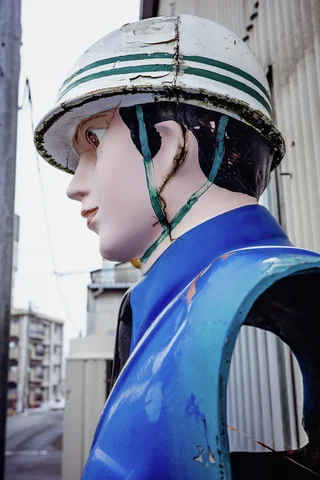 A weathered painted figure in blue wears a cracked white helmet with green stripes and chin straps.