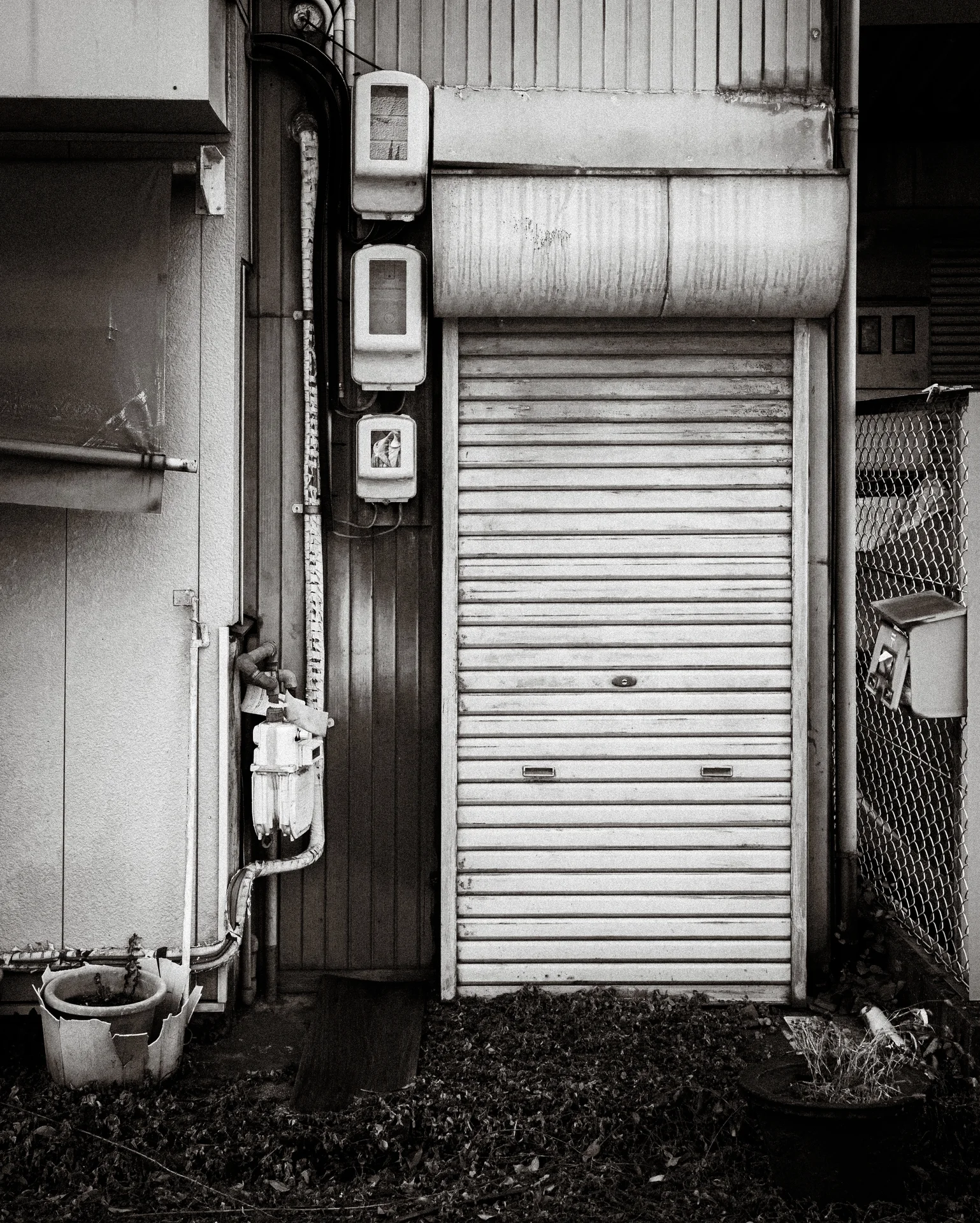 A narrow closed metal shutter stands beside exposed pipes and small wall fixtures in black and white.