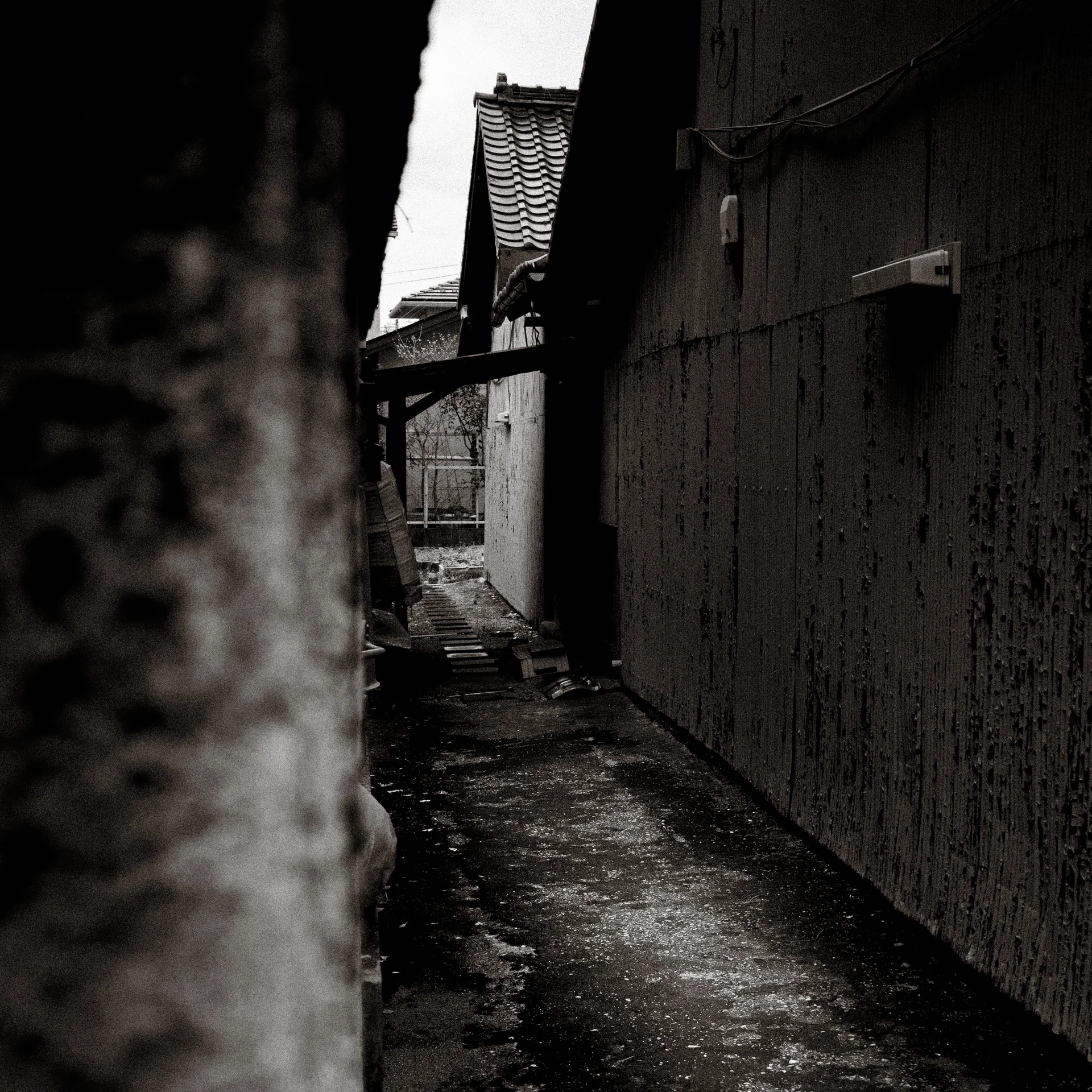 A narrow uneven path runs between dark weathered walls beneath projecting tiled roofs in black and white.