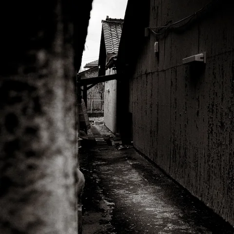 A narrow uneven path runs between dark weathered walls beneath projecting tiled roofs in black and white.