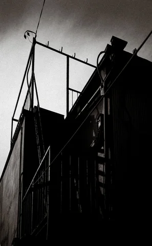 A skeletal rectangular metal frame rises above a dark building beneath cloudy sky in black and white.