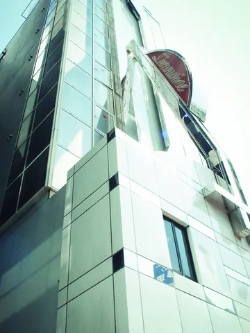 A pale tiled building rises steeply beside reflective glass windows and a projecting oval rooftop sign.