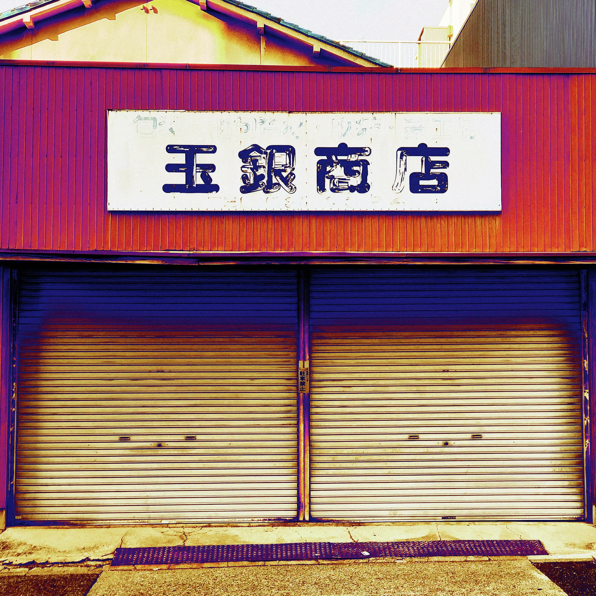 A white Japanese sign hangs above two closed cream shutters on a reddish corrugated storefront.