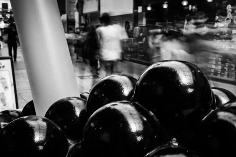 Glossy black rounded forms dominate the foreground as blurred shoppers pass behind a white column.