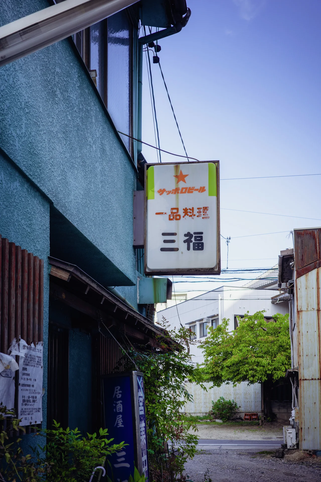 A rectangular hanging sign projects from a blue building above a narrow passage ending at leafy trees.