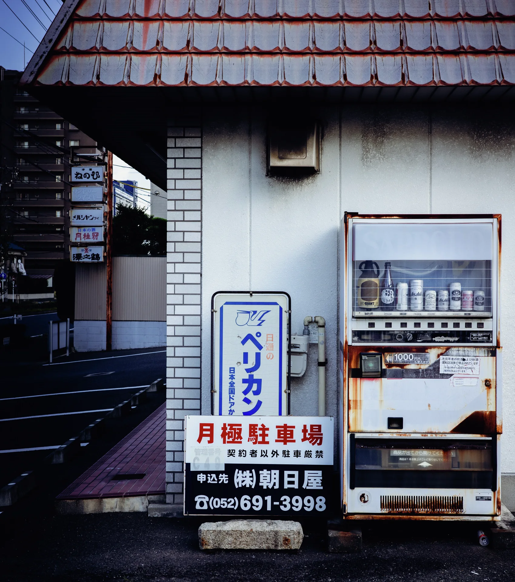 A rusted vending machine stands beside parking signs against a pale wall beneath tiled eaves.