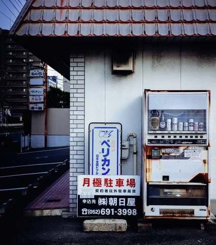 A rusted vending machine stands beside parking signs against a pale wall beneath tiled eaves.