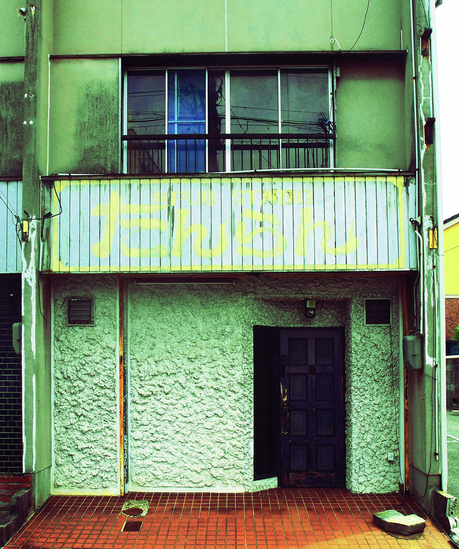 A faded striped sign spans a narrow green building above a dark doorway and textured pale wall.