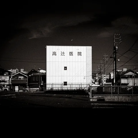 A bright rectangular building stands among low rooftops and utility poles beneath a dark sky.