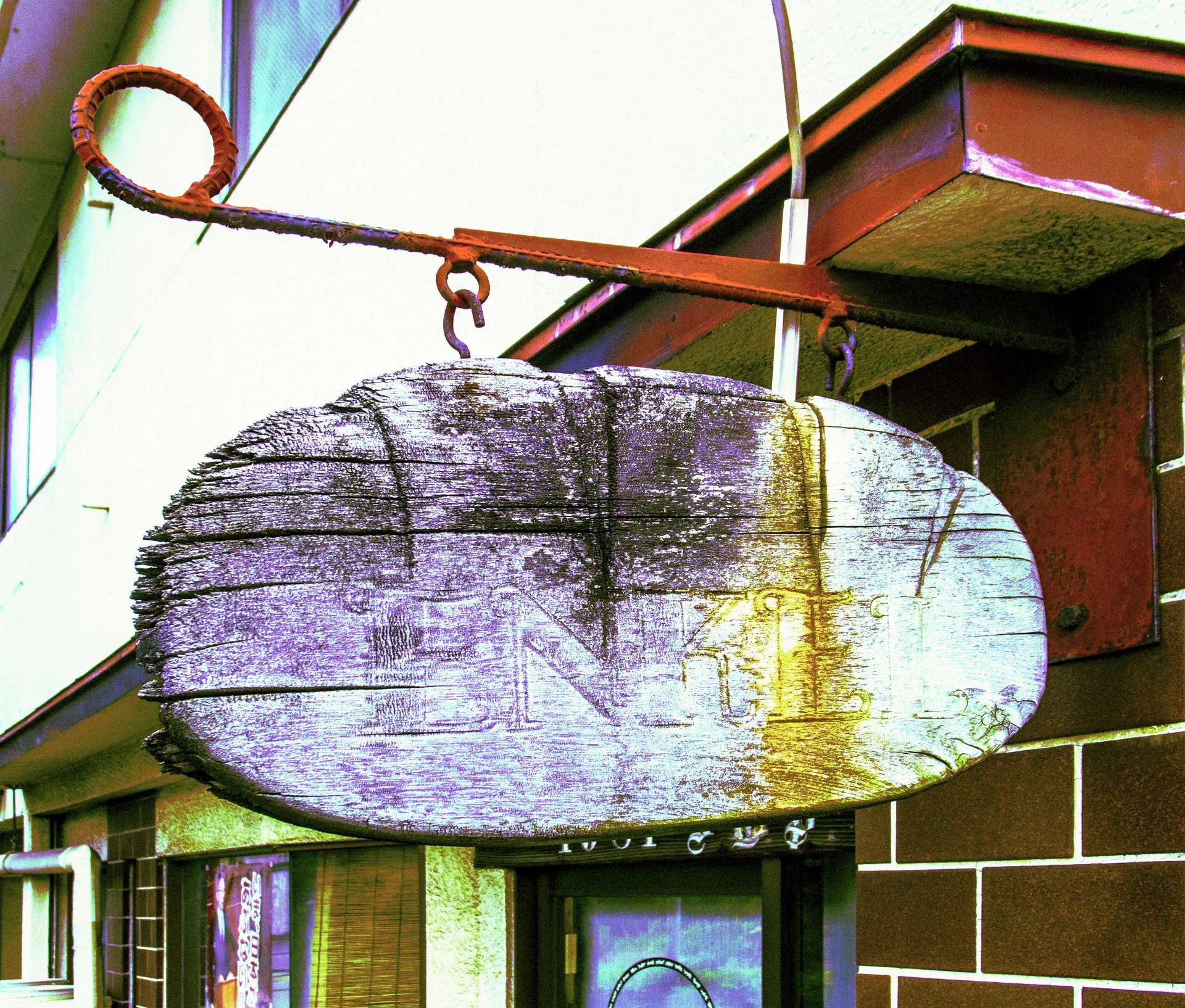 A faded oval wooden sign hangs from a rusty bracket outside a brown tiled building.