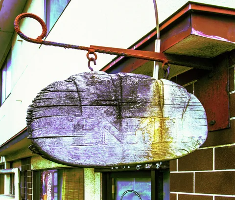 A faded oval wooden sign hangs from a rusty bracket outside a brown tiled building.