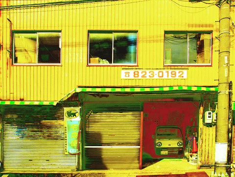 A small car sits behind a partly raised shutter beneath yellow corrugated siding and three upper windows.