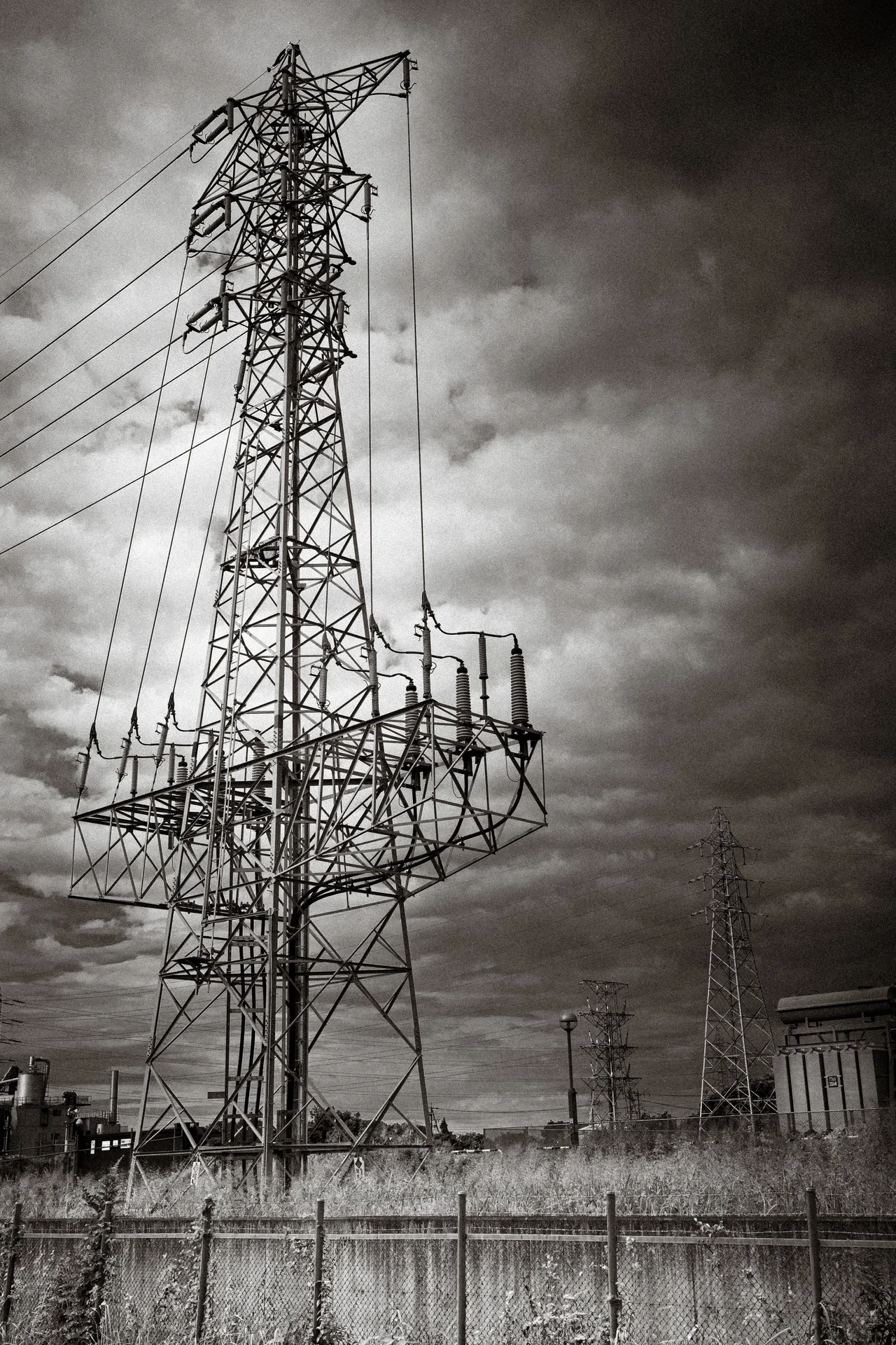 A tall lattice electricity pylon rises behind a concrete fence beneath textured clouds in black and white.
